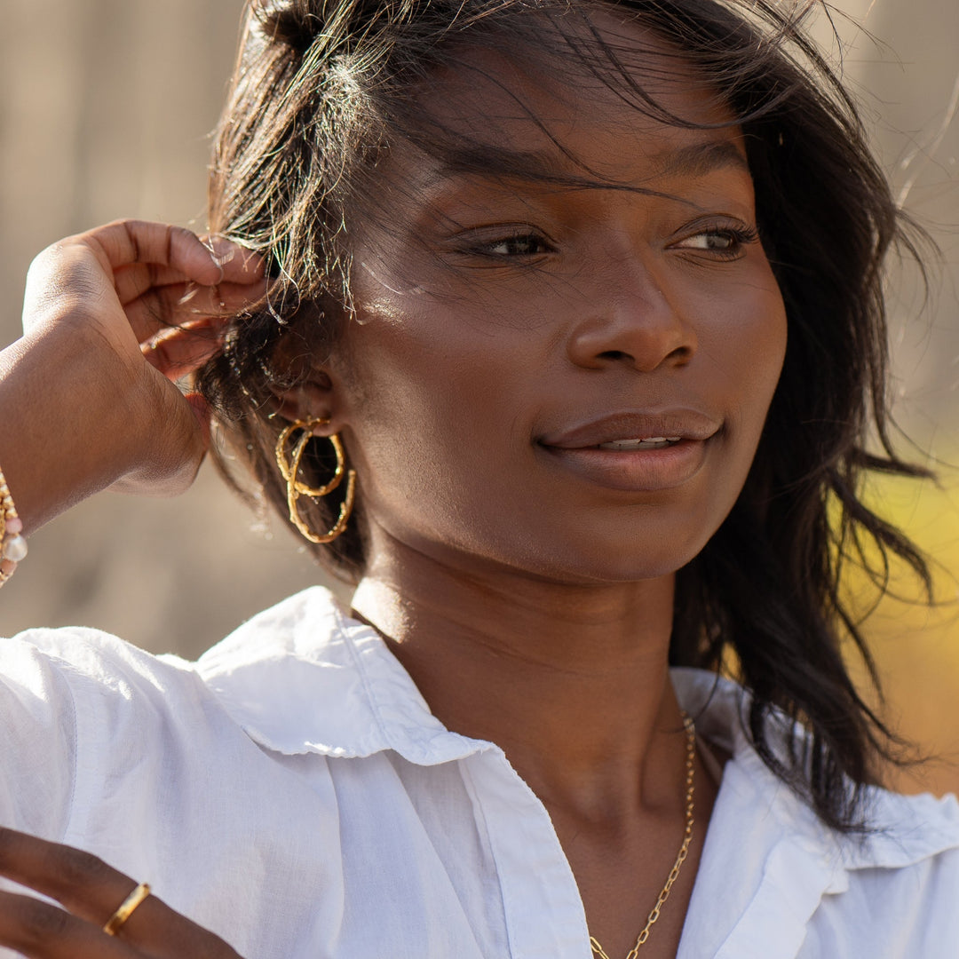 Close-up of a woman wearing elegant gold hoop earrings, layered gold necklaces, and a white blouse, styled with a minimalistic and natural look. Wind-swept hair and soft lighting enhance the earthy and boho vibe.