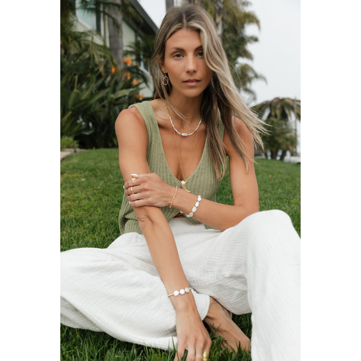 Boho-chic woman with long blonde hair sitting on lush green grass, wearing layered gold and pearl jewelry, including a pearl bracelet, gold rings, and a delicate layered necklace, exuding effortless coastal style.