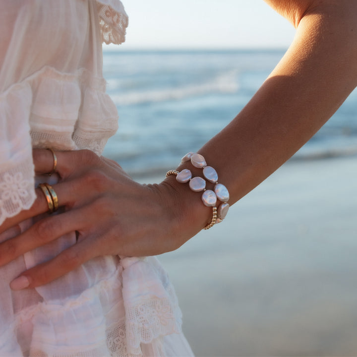 Close-up of a woman's wrist adorned with two gold beaded bracelets featuring lustrous freshwater pearl coins, set against a serene beach backdrop with ocean waves.