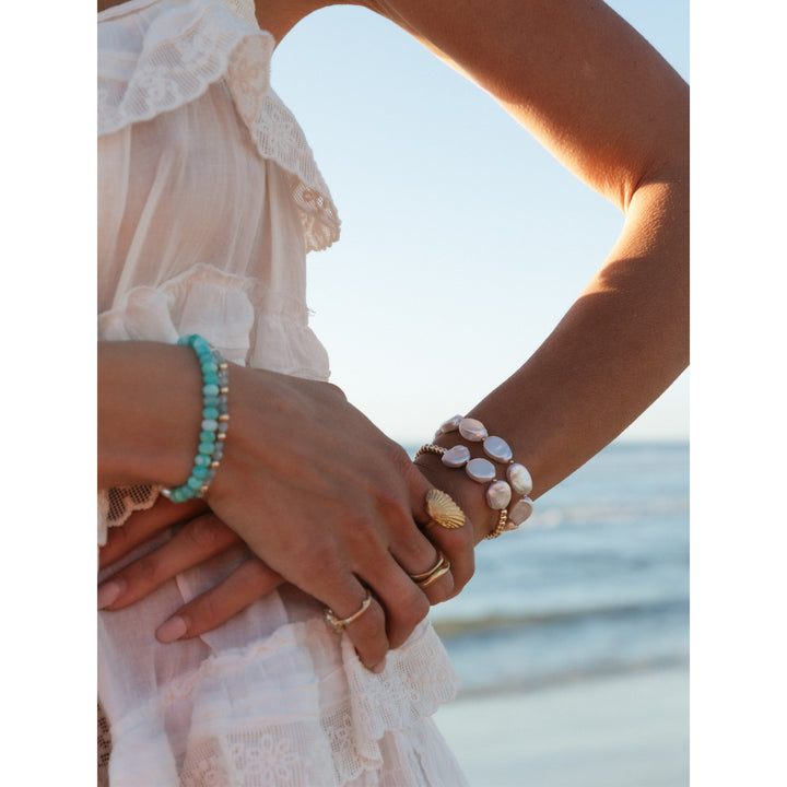 Close-up of a woman's hands adorned with gold rings, a Amazonite beaded bracelet, and two gold beaded bracelets with freshwater pearl coins, set against a serene beach backdrop with ocean waves.