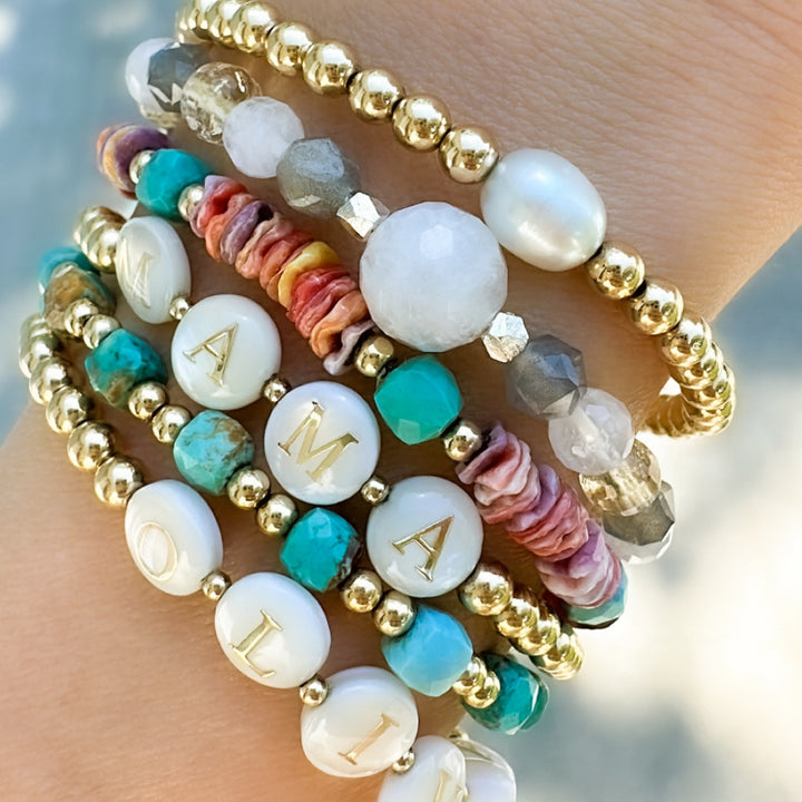 Close-up of a wrist adorned with stacked gold beaded bracelets featuring colorful gemstones, freshwater pearls, shell accents, and personalized alphabet beads spelling 'MAMA' and 'OLIVIA,' creating a vibrant and meaningful jewelry stack.