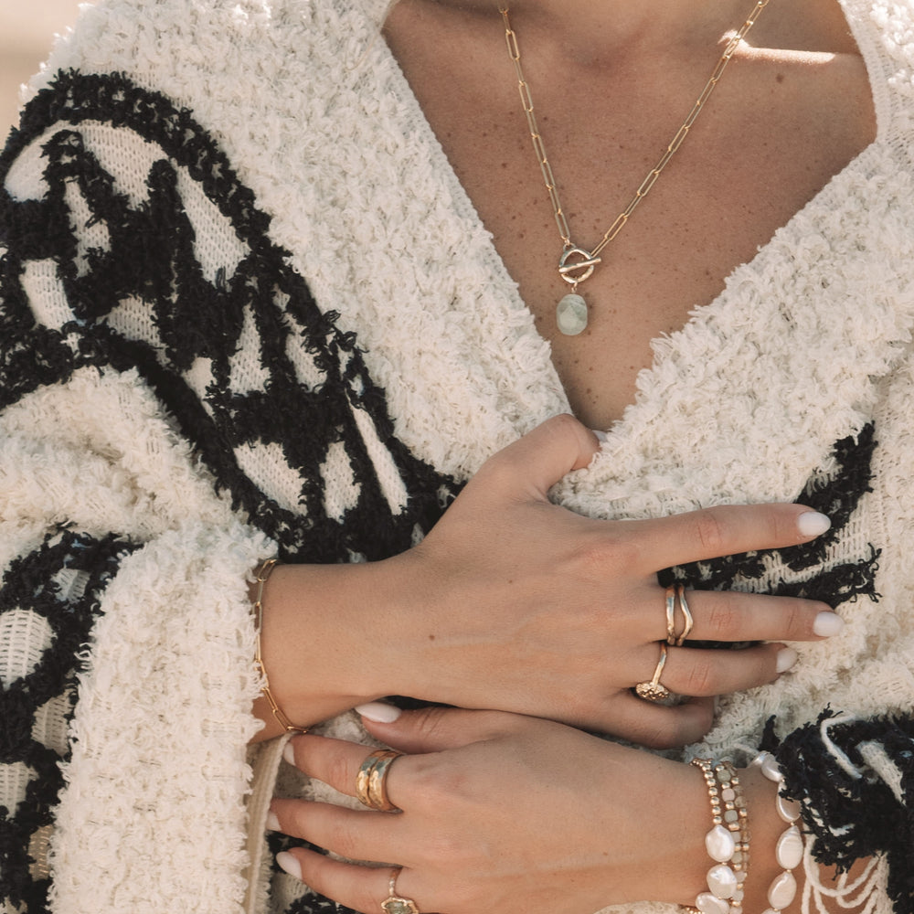Close-up of a woman wearing the Inner Calm Aquamarine Toggle Necklace with a hand-sculpted gold toggle clasp on a paperclip chain. Styled with gold rings, layered bracelets, and a cozy cream and black textured wrap, evoking a serene coastal boho vibe, by Blooming Lotus Jewelry.