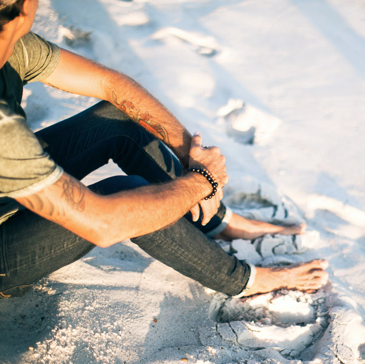 man sitting on beach wearing onyx and sandalwood wrist malas - blooming lotus jewelry