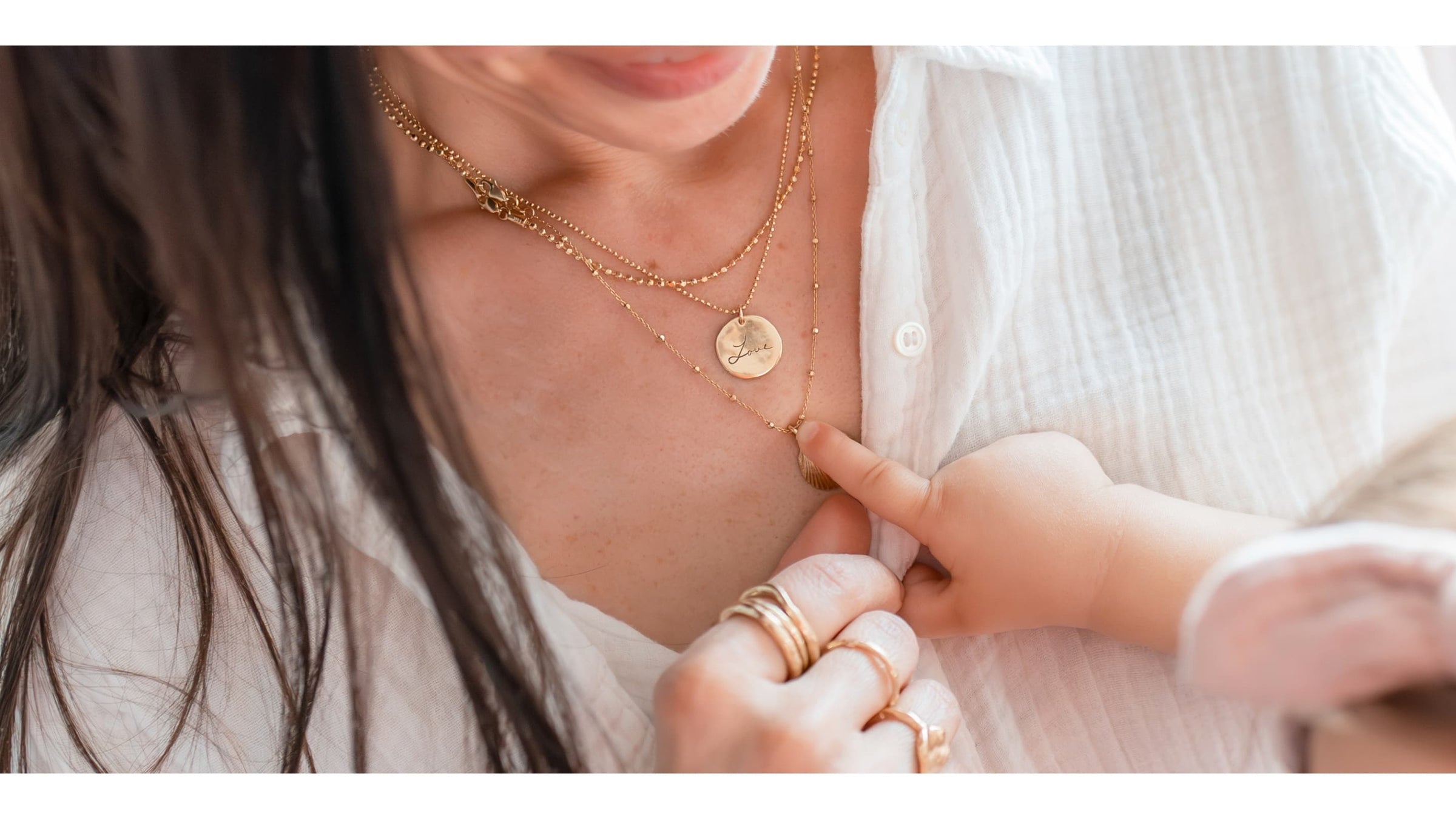 Close-up of a woman wearing layered gold necklaces featuring a personalized engraved pendant with the word “Love.” A baby’s hand gently touches the necklace. Handcrafted jewelry by Blooming Lotus Jewelry.