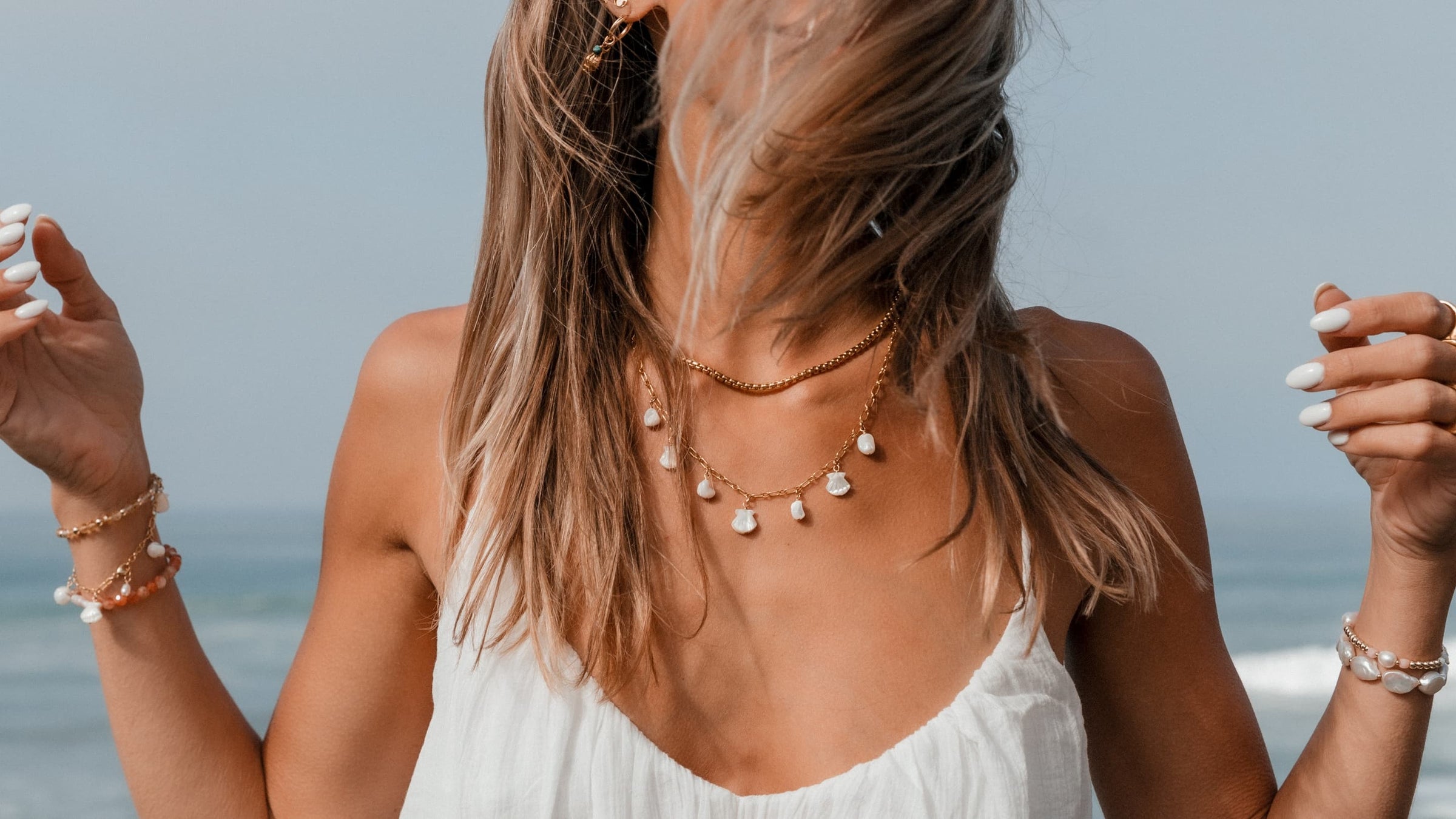 Woman wearing Blooming Lotus Jewelry at the beach, layered in gold chain necklaces with shell and pearl charms, and coastal gemstone bracelets. Handmade boho jewelry for ocean lovers.