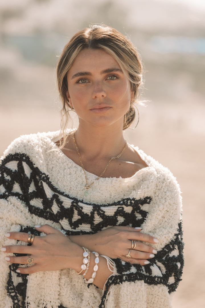 Woman standing on the beach wearing Blooming Lotus Jewelry’s Inner Calm Aquamarine Toggle Necklace with a hand-carved gold toggle clasp on a paperclip chain. Styled with gold rings, pearl and gemstone bracelets, and a cozy cream and black textured wrap. The soft sunlight highlights the soothing blue-green aquamarine gemstone, capturing a calm and coastal boho vibe.