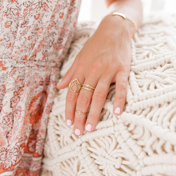 Close-up of a hand resting on a macrame cushion, adorned with layered gold rings including the Lotus Ring, the Tiny Eye of Protection Ring, and a textured Olive Branch Twig stacking ring. The intricate designs complement the bohemian floral dress, creating an elegant and effortless look. A gold bracelet adds a final touch of sophistication to the styling.