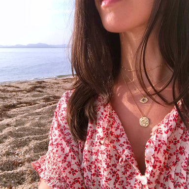 Gold Om Necklace and Evil Eye Necklace on model wearing white and red top at beach - Blooming Lotus Jewelry