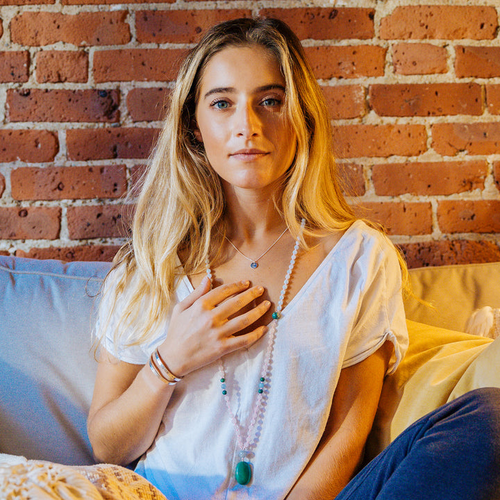 Serene woman seated on a cozy couch with a brick wall backdrop, wearing a minimalist silver necklace with a tiny round charm, complemented by a long mala beads necklace featuring rose quartz and a Turquoise pendant. Her relaxed, natural look embodies a peaceful and mindful yoga lifestyle.