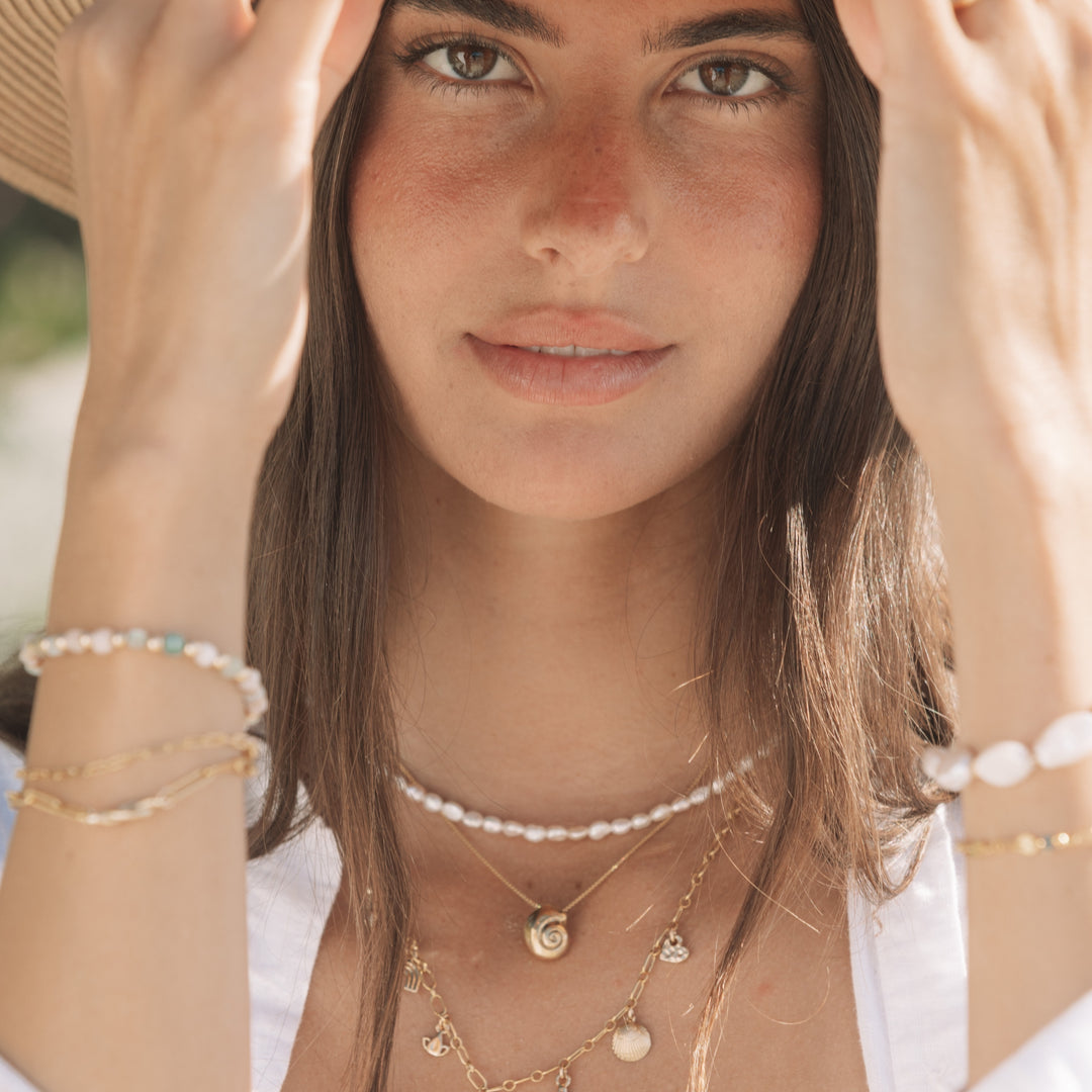 Woman wearing the Becoming Necklace featuring a gold spiral shell pendant layered with pearl and charm necklaces, styled in soft coastal light by Blooming Lotus Jewelry.