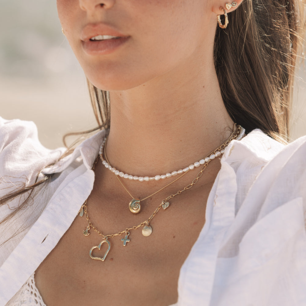 Close-up of woman wearing the Becoming Necklace (a gold spiral shell pendant)  layered with a freshwater pearl choker and gold charm necklace, styled in soft coastal light by Blooming Lotus Jewelry.