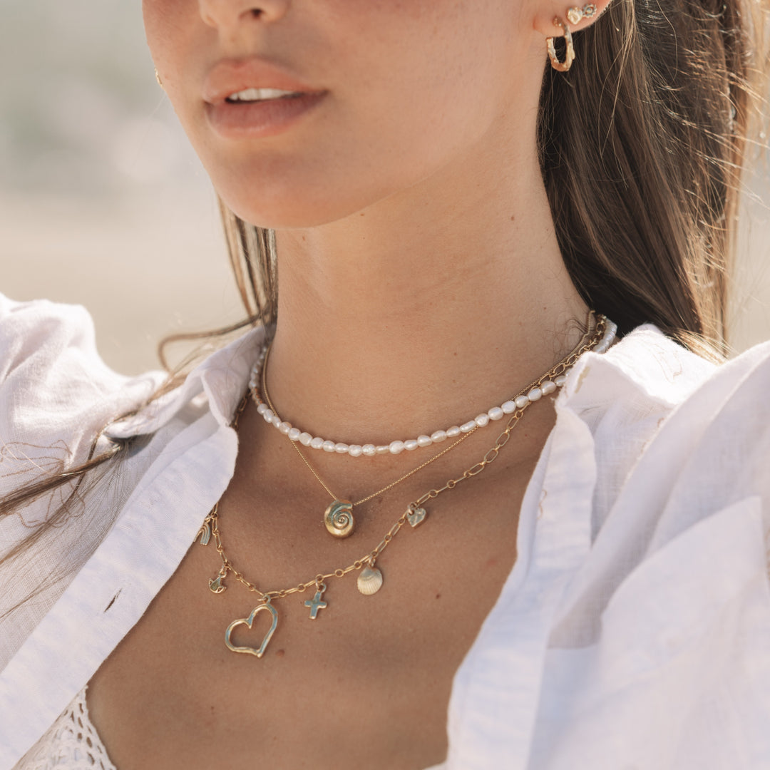 Close-up of woman wearing the Becoming Necklace (a gold spiral shell pendant)  layered with a freshwater pearl choker and gold charm necklace, styled in soft coastal light by Blooming Lotus Jewelry.