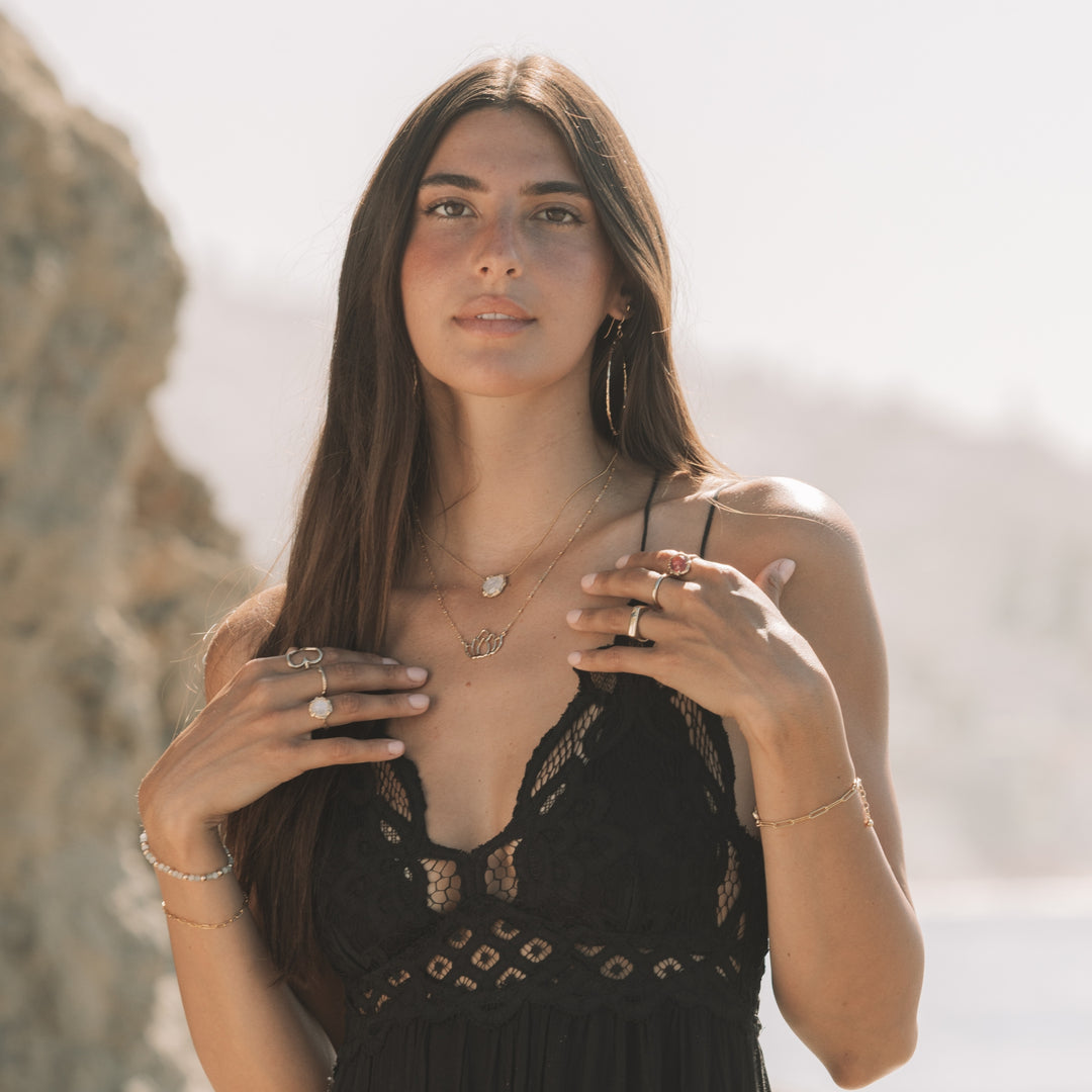 Model standing by the coast wearing layered Blooming Lotus Jewelry pieces, including a gold lotus necklace, moonstone necklace, gold hoops, and gemstone rings. A soft, yoga inspired jewelry look styled with a black lace dress in natural sunlight.