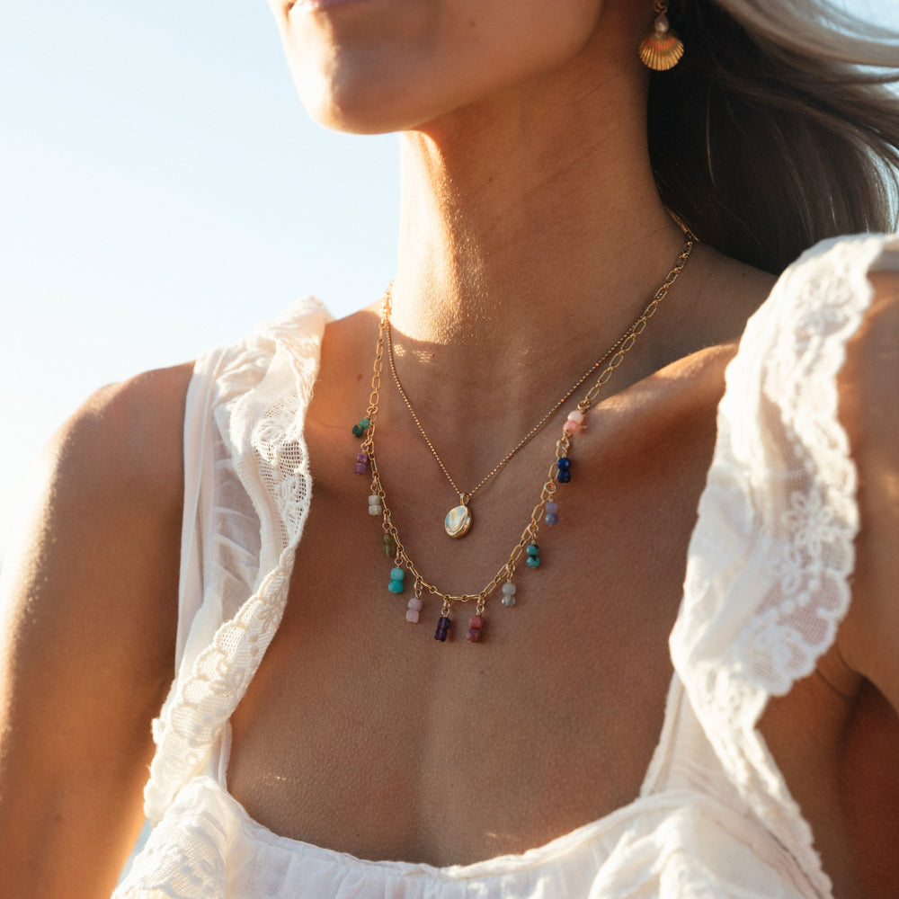 Close-up of a woman wearing a white lace-trimmed dress, layered with two gold necklaces — one featuring a coin Fingerprint pendant and the other adorned with colorful gemstone charms by Blooming Lotus Jewelry. She is also wearing a gold shell earring, with golden light from the setting sun illuminating the scene.