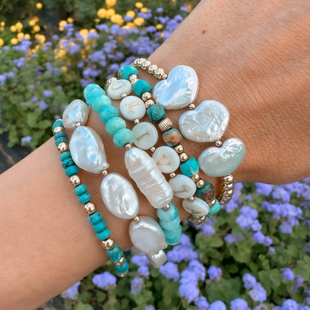 Stack of Blooming Lotus Jewelry bracelets featuring turquoise beads, freshwater pearls, gold accents, and heart-shaped pearls, styled on wrist with purple and yellow flowers in background.