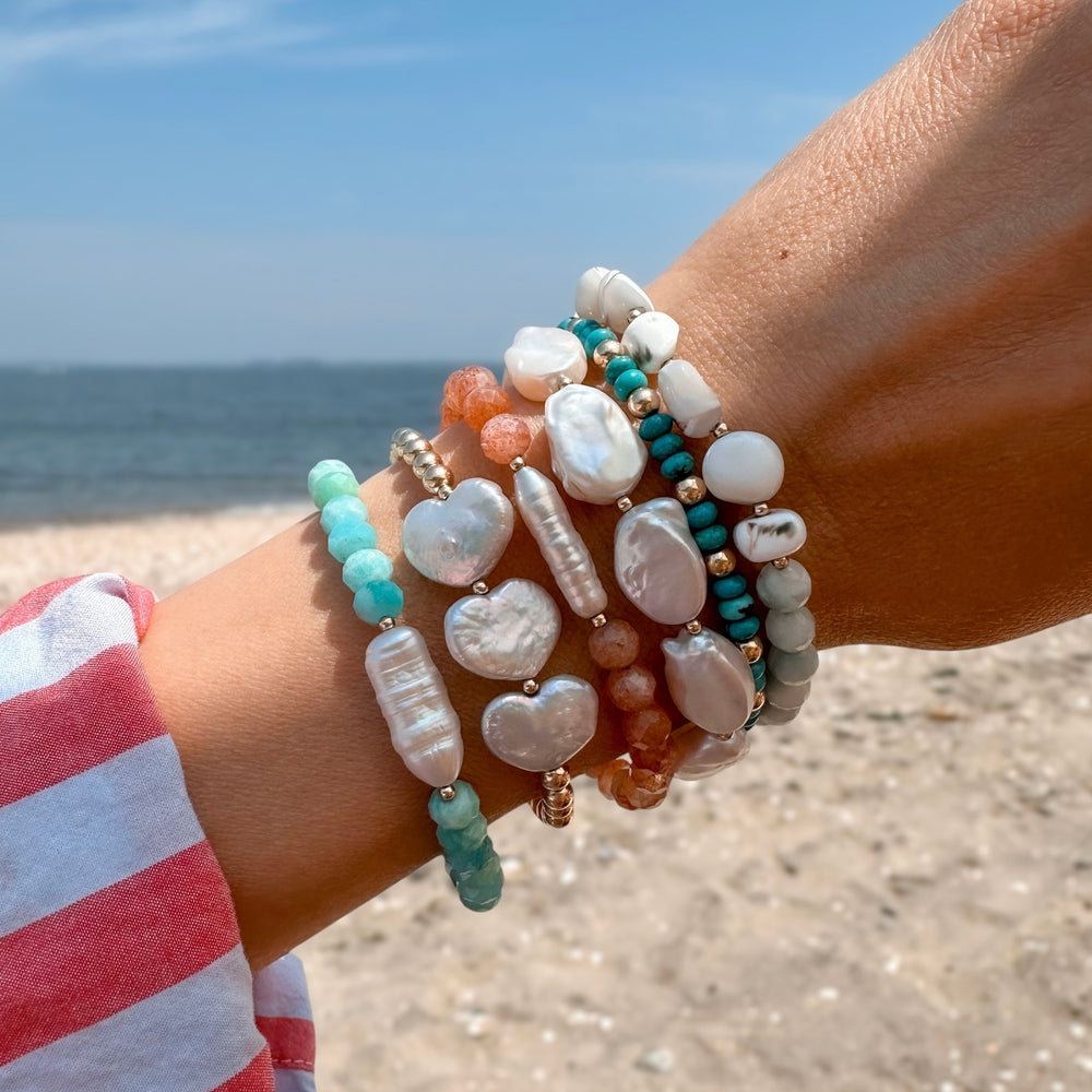 Close-up of a wrist stacked with coastal-inspired gemstone and freshwater pearl bracelets, including the Sunkissed and Seafoam Serenity Bracelets, against a sandy beach and ocean backdrop.