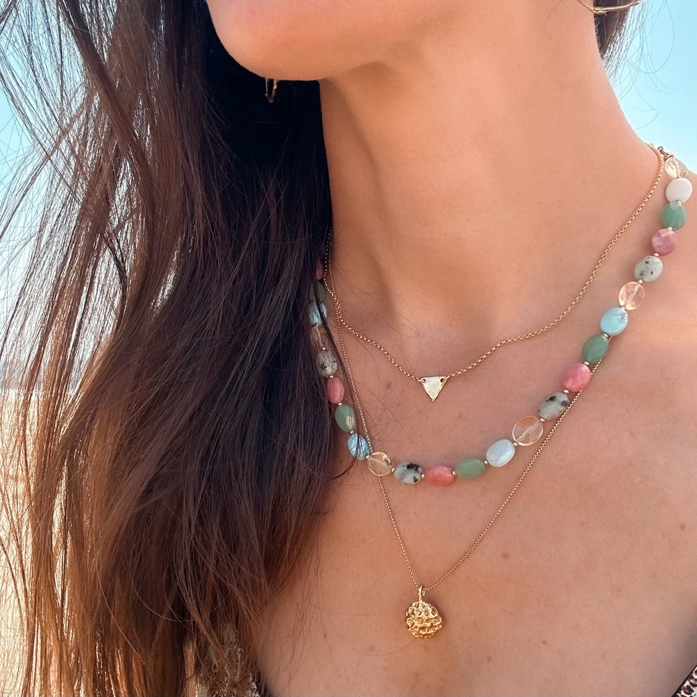 Woman wearing the Coral Cove Necklace, a strand of multi-gemstone beads in pink, green, aqua, yellow, and gray with gold accents, styled for a colorful, coastal boho-inspired look by Blooming Lotus Jewelry. It is paired with the Coral Bloom Necklace and inverted triangle necklace.
