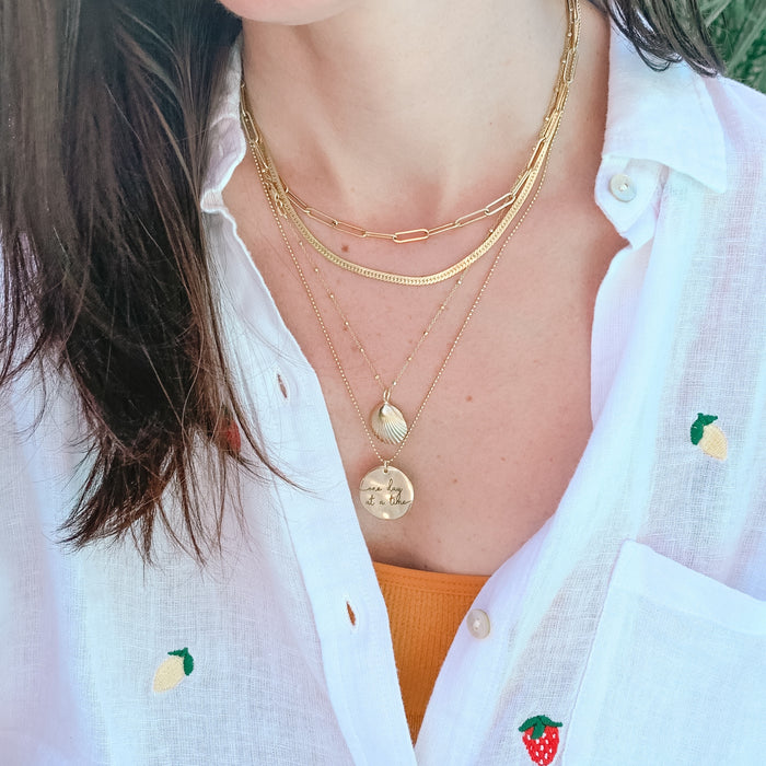 Model wearing gold layering necklace paperclip, herringbone, large seashell necklace 18 inches, mantra coin 20 inches