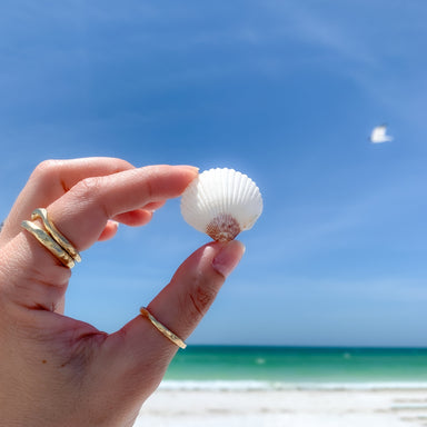 gold stacking rings on womans fingers while holding a seashell at the beach with ocean and blue sky