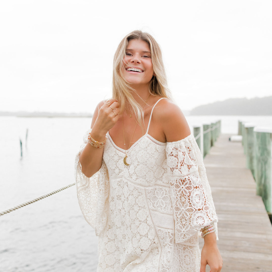 A smiling woman standing on a dock by the water, wearing a bohemian-style white lace dress and accessorized with the Luna Crescent Moon Necklace, layered gold jewelry, and colorful gemstone bracelets. Her hair flows naturally in the breeze, complementing the serene, coastal backdrop.