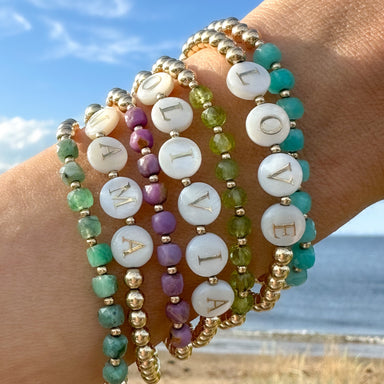 gold beaded bracelets with gemstones and personalized white alphabet beads on models wrist with beach in background