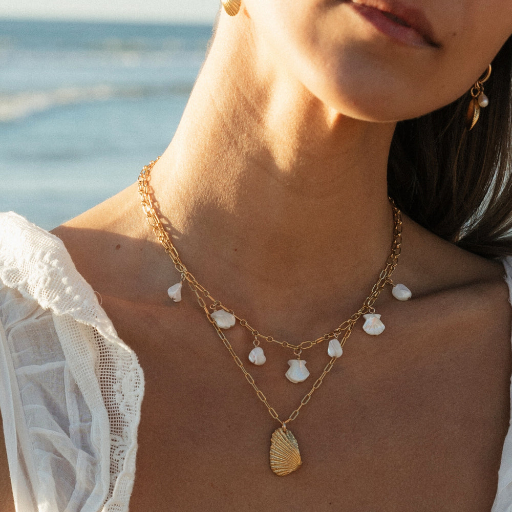 Close-up of a woman wearing two layered gold necklaces from Blooming Lotus Jewelry — one with dangling white mother-of-pearl shell charms and the other with a textured gold seashell pendant. She is dressed in a white lace-trimmed top, with golden sunlight and the ocean softly blurred in the background.