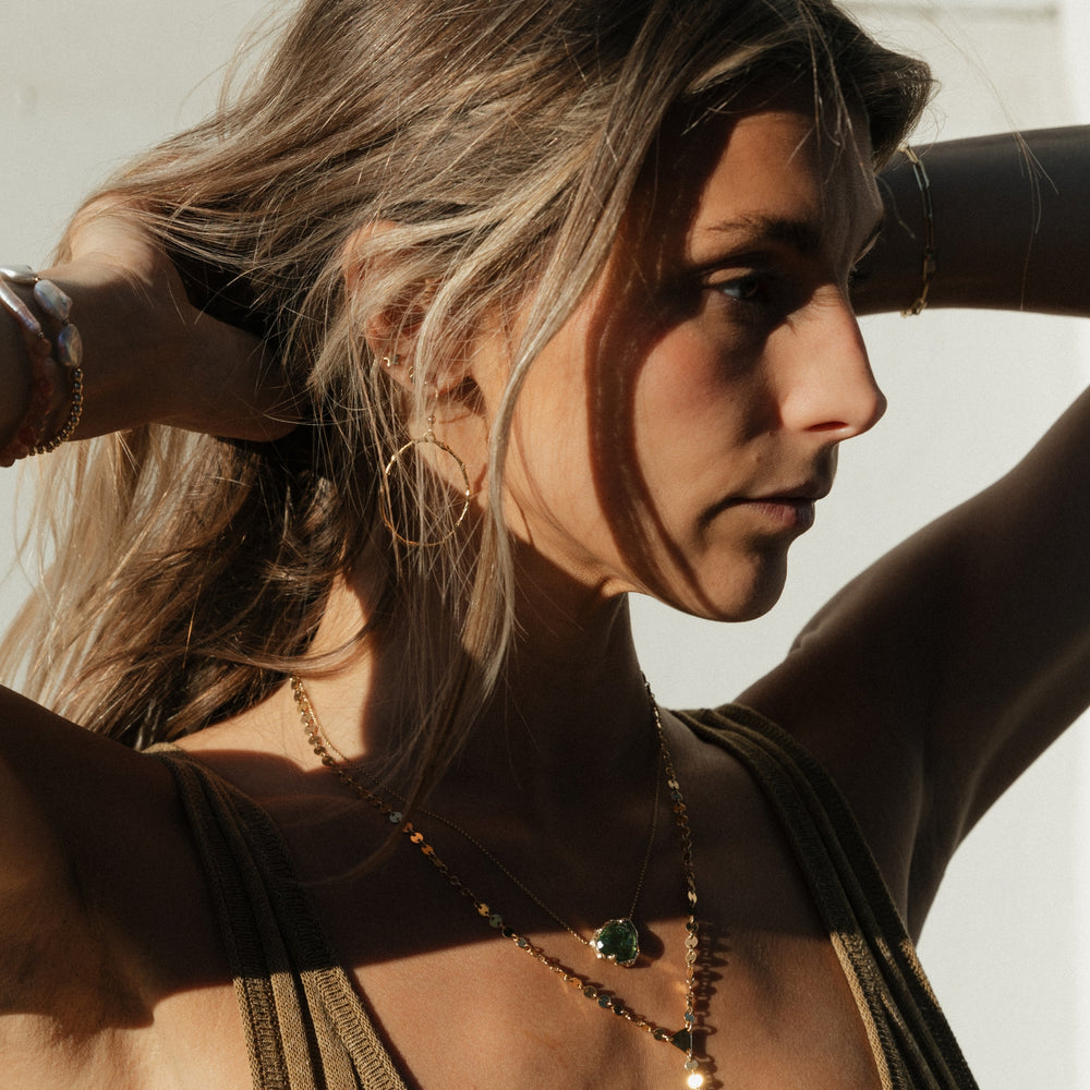 Close-up of a woman with sun-kissed skin wearing Blooming Lotus Jewelry’s Olive Branch Loop Hoops, featuring delicate gold branch-like texture, along with layered gold necklaces and bracelets in natural light. Model: Taylor Nicole Hayden