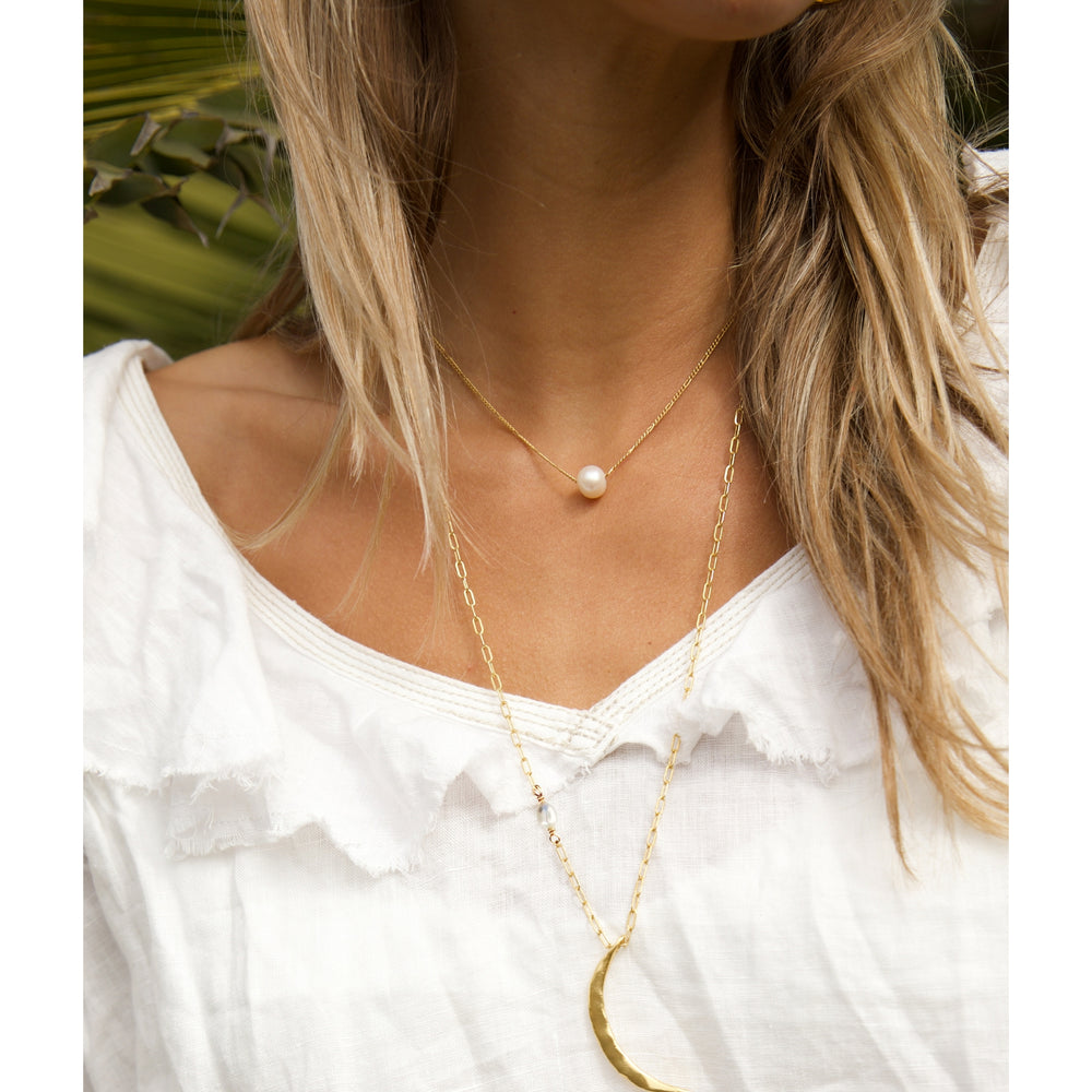 Close-up of a woman wearing the Pearl Drift Necklace by Blooming Lotus Jewelry, featuring a single pearl that slides freely on a delicate gold Figaro chain, layered with a long skinny crescent moon necklace over a white ruffled linen blouse.