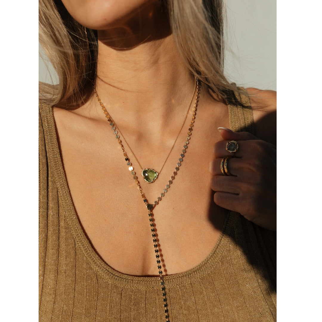 Close-up of woman wearing the Sol Lariat Necklace layered with a green tourmaline pendant. The gold disc chain catches the sunlight, adding a touch of sparkle to her neutral-toned outfit.
