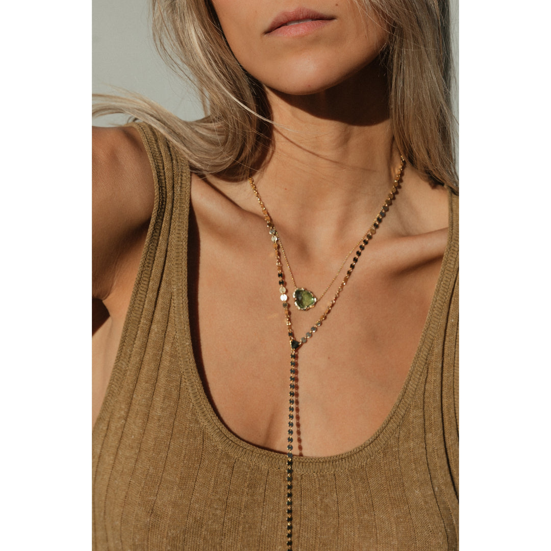 Close-up of woman wearing the Sol Lariat Necklace layered with a green tourmaline pendant. The gold chain catches the light beautifully against her tan ribbed tank and sun-kissed skin.