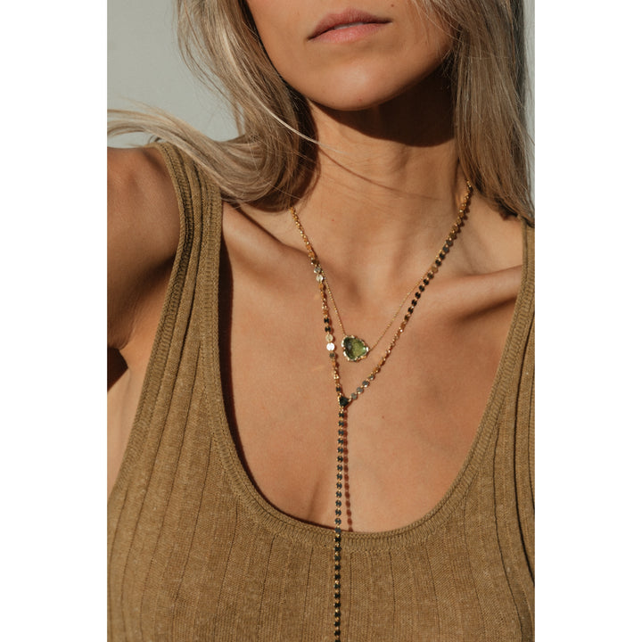 Close-up of woman wearing the Sol Lariat Necklace layered with a green tourmaline pendant. The gold chain catches the light beautifully against her tan ribbed tank and sun-kissed skin.