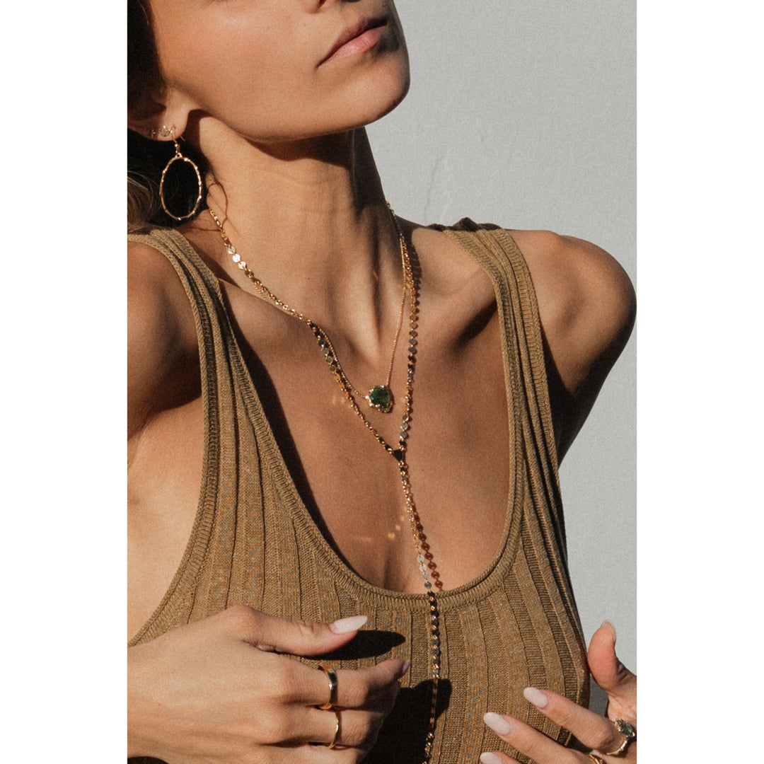 Sun-kissed model wearing Blooming Lotus Jewelry, featuring the gold SOL Lariat Necklace layered with a green tourmaline pendant necklace and large branch gold hoops. A boho, coastal-inspired look with delicate rings and golden hour lighting.