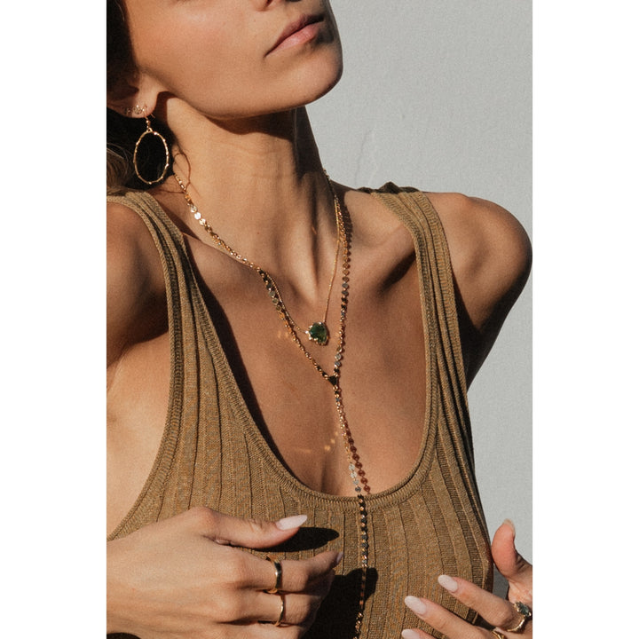 Sun-kissed model wearing Blooming Lotus Jewelry, featuring the gold SOL Lariat Necklace layered with a green tourmaline pendant necklace and large branch gold hoops. A boho, coastal-inspired look with delicate rings and golden hour lighting.