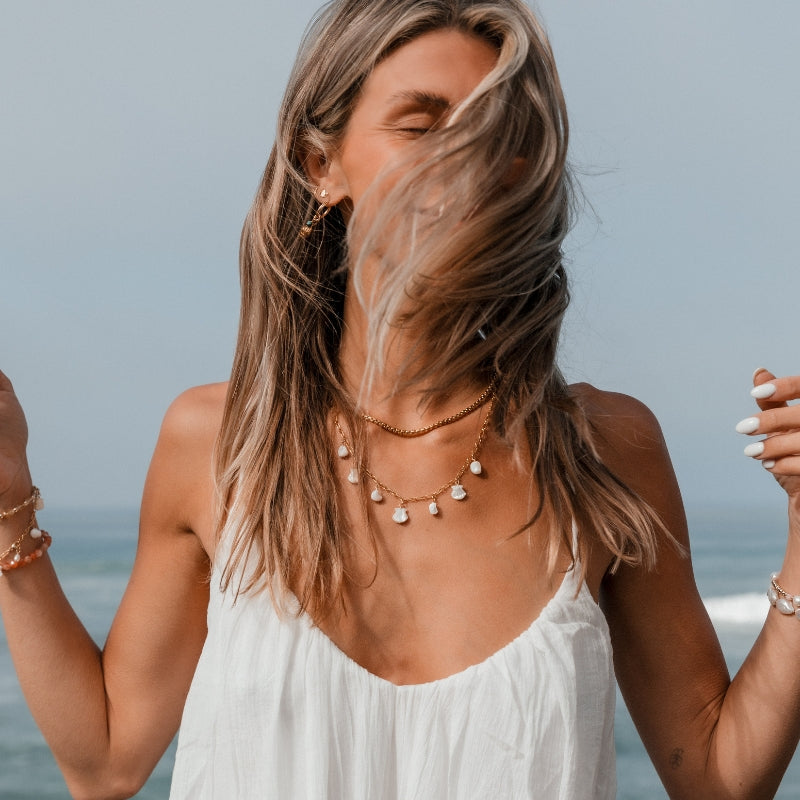 Woman wearing Blooming Lotus Jewelry at the beach, layered in gold chain necklaces with pearl and shell charms, gold shell hoop earrings and studs, and coastal-inspired gemstone bracelets. Handcrafted boho jewelry for ocean lovers.