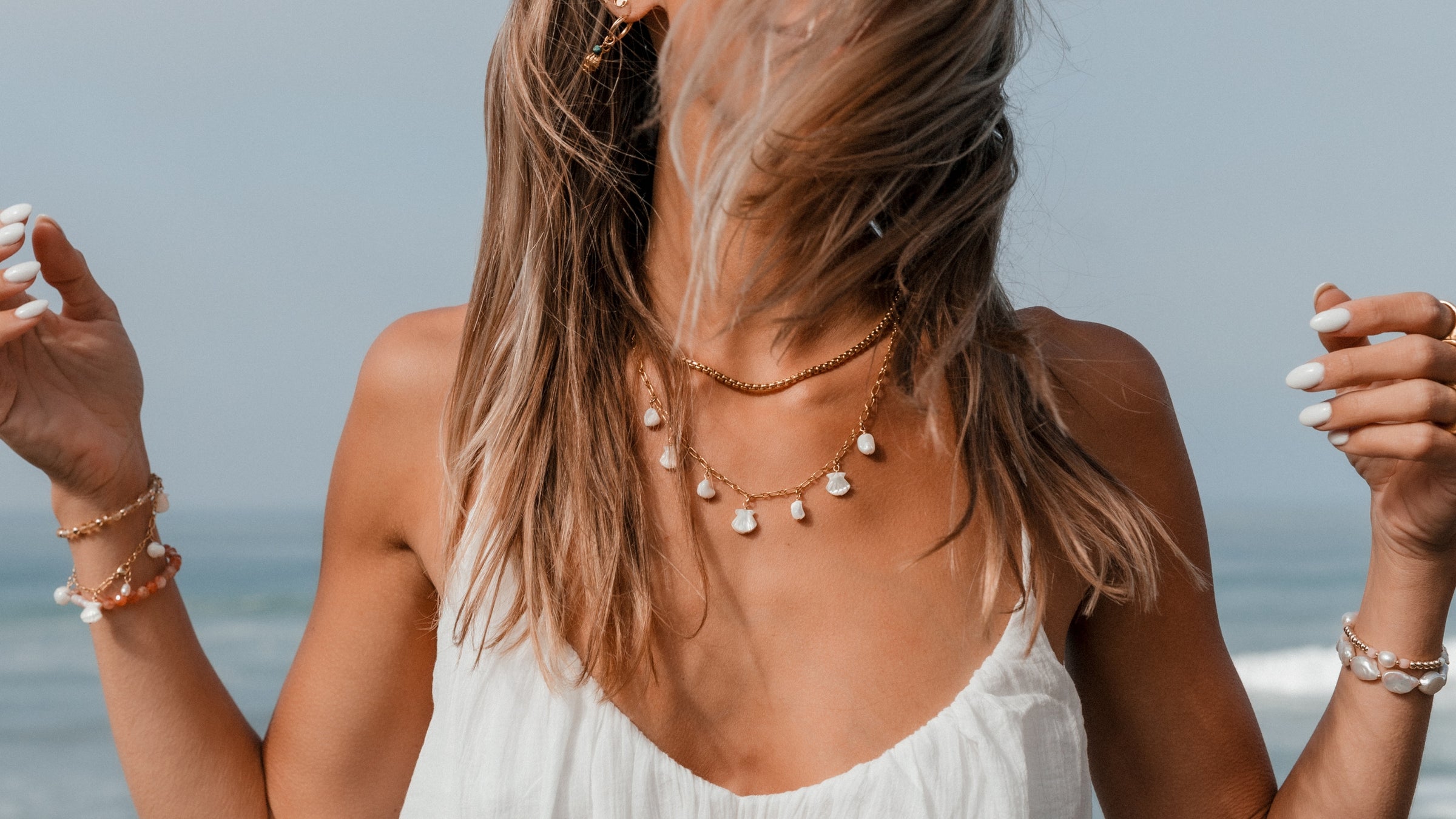 Woman wearing Blooming Lotus Jewelry at the beach, layered in gold chain necklaces with pearl and shell charms, and coastal-inspired gemstone bracelets. Handcrafted boho jewelry for ocean lovers.