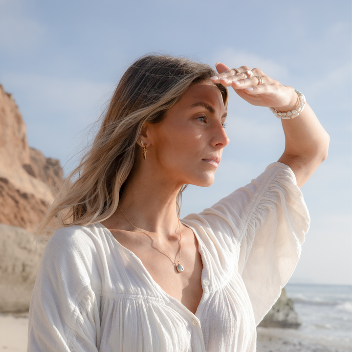 Woman standing on a sunny beach wearing coastal boho gold jewelry, gently shading her eyes as she looks toward the ocean.