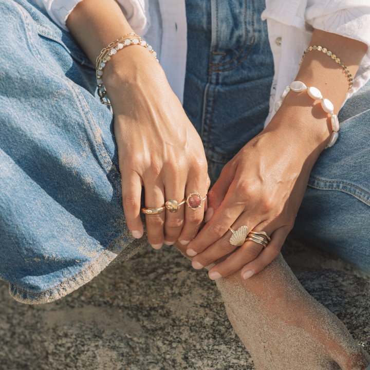 Close-up of hands resting on sand and denim, wearing stacked gold rings including a seashell ring, a Pink Tourmaline ring, with layered beaded and gold bracelets in a coastal beach setting.