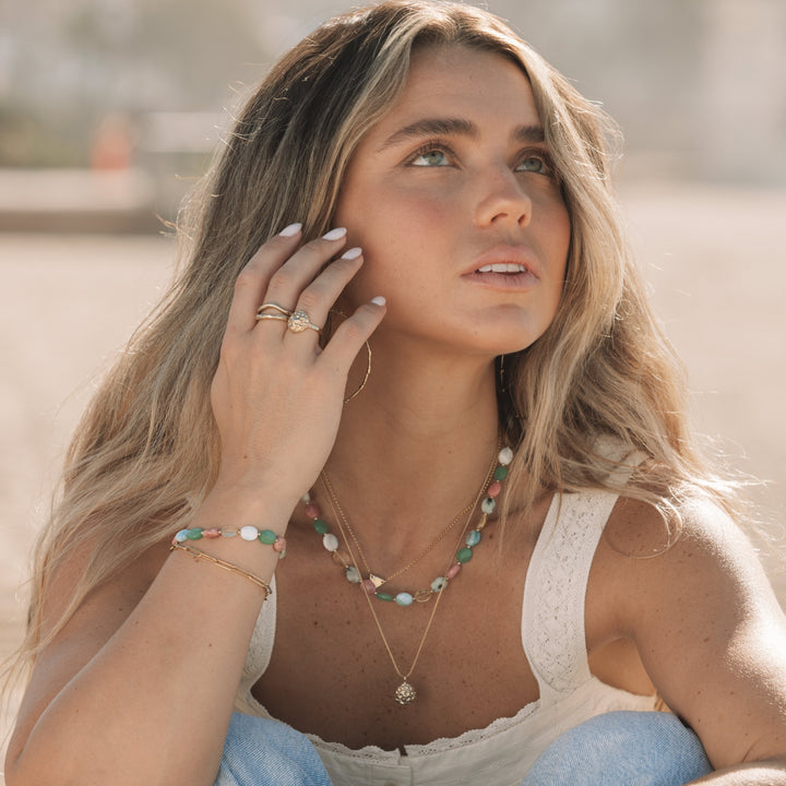 woman sitting on the beach wearing the coral bloom ring and matching gold pendant necklace, layered gemstone necklaces and bracelets, coastal boho jewelry by Blooming Lotus Jewelry