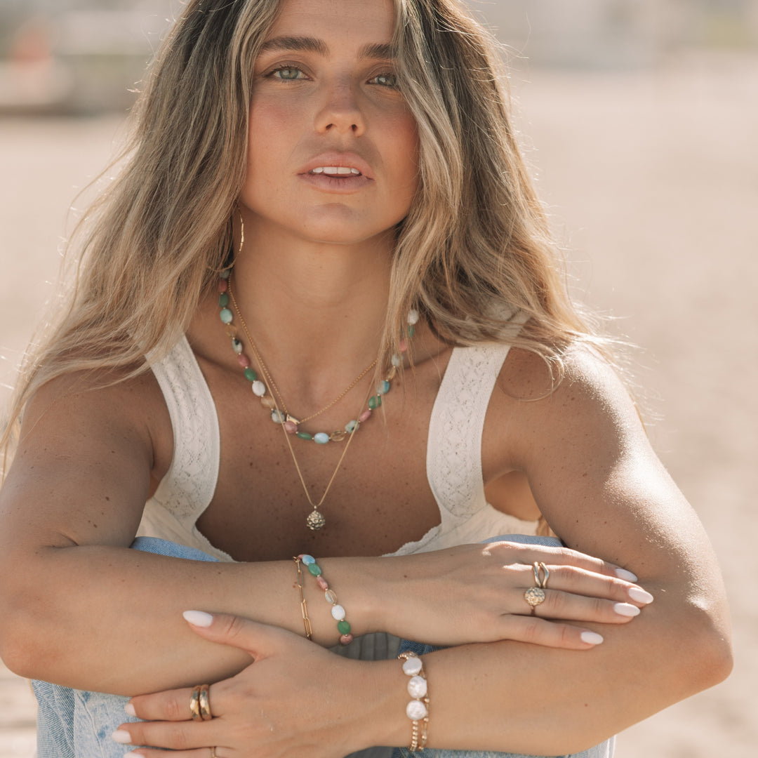 woman sitting on the beach wearing the coral bloom ring and matching gold pendant necklace, layered gemstone necklaces and bracelets, coastal boho jewelry by Blooming Lotus Jewelry