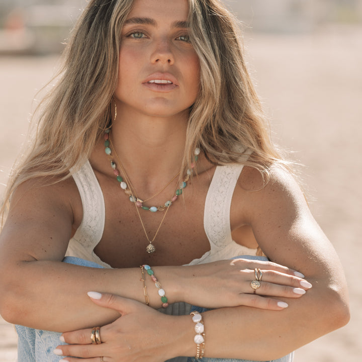 woman sitting on the beach wearing the coral bloom ring and matching gold pendant necklace, layered gemstone necklaces and bracelets, coastal boho jewelry by Blooming Lotus Jewelry