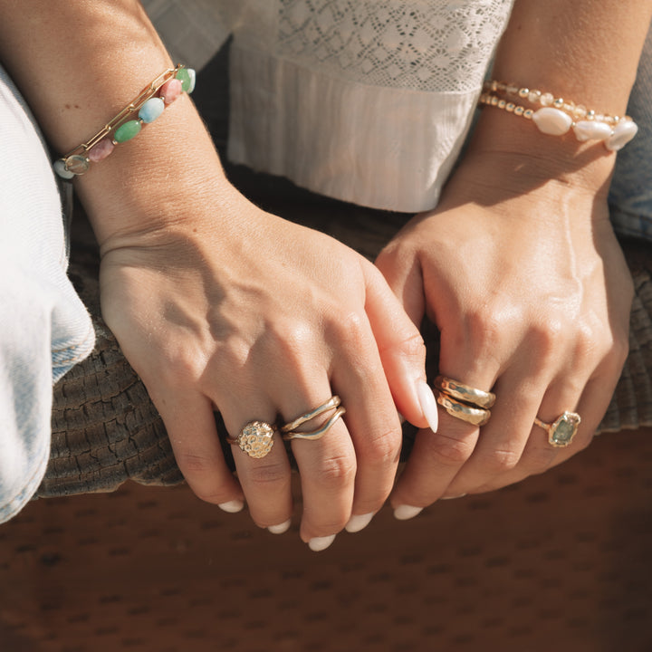 Woman's hands resting on a wood bench, wearing the Coral Bloom Ring stacked with two dome rings, gold stacking bands, paired with gemstone and pearl bracelets.