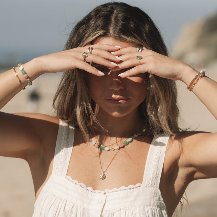 woman on the beach wearing the coral bloom necklace and matching gold rings, shielding her eyes from the sun, coastal boho jewelry by Blooming Lotus Jewelry