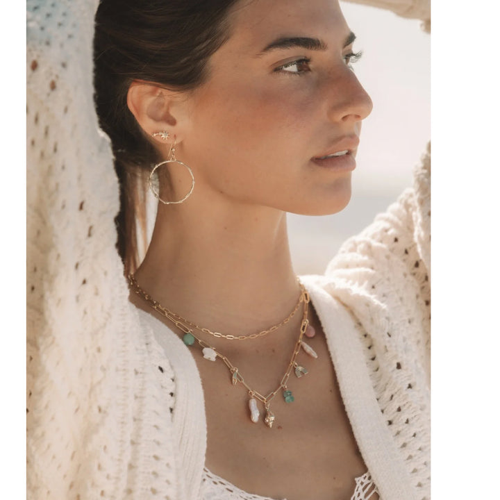 Lifestyle image of a woman wearing the Blooming Lotus Jewelry Custom Charm Necklace layered with gold chains, gold branch hoops, and studs, styled with a cream sweater for a boho coastal look.