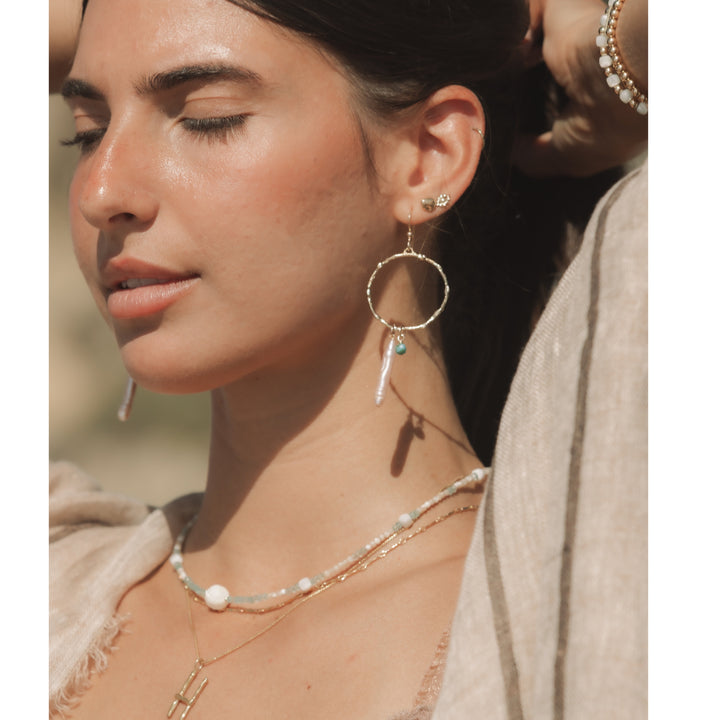 Woman with eyes closed wearing gold Branch Hoops with turquoise cube and Biwa pearl dangle, layered gold large initial necklace and gemstone necklace, and neutral linen top in soft beach sunlight.