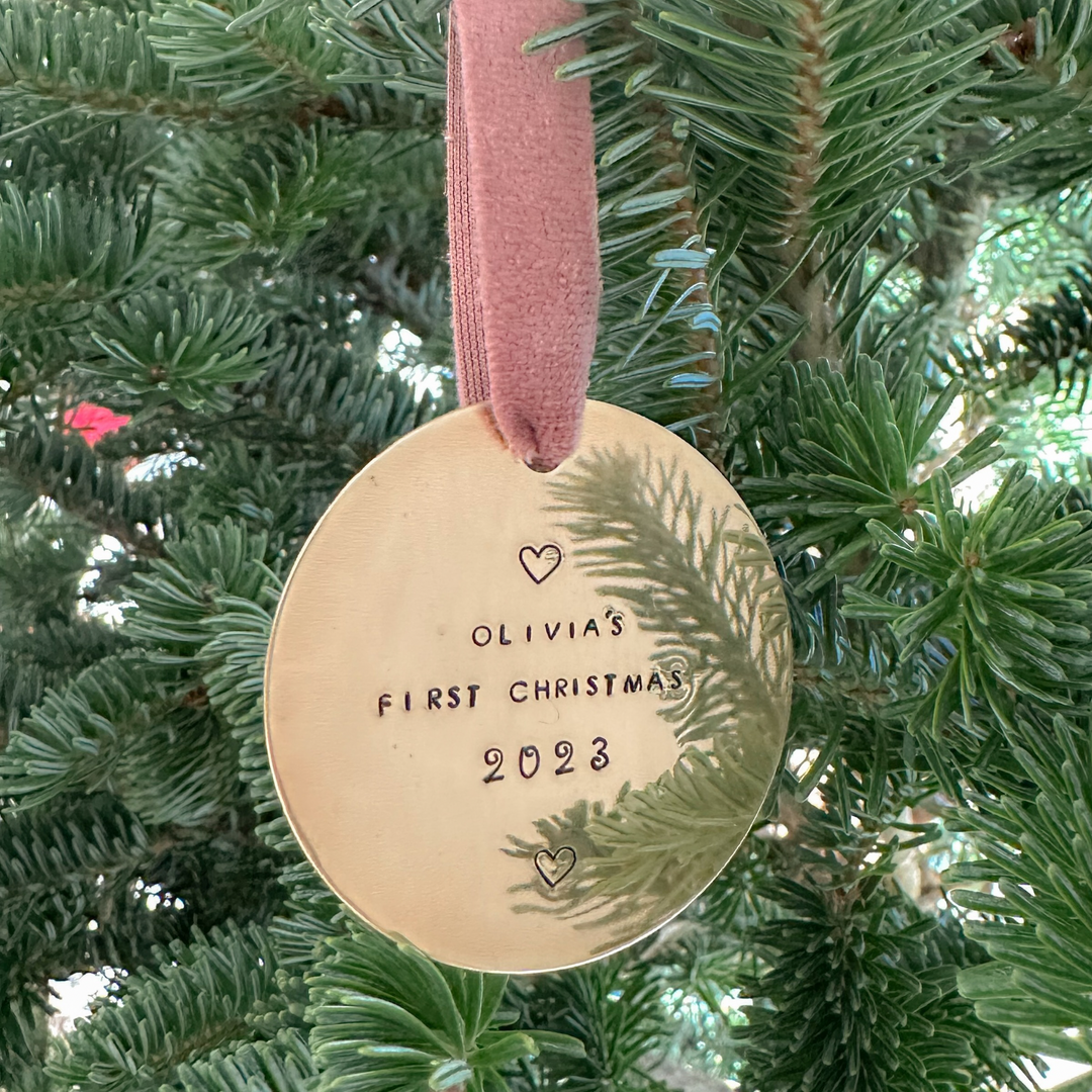Gold round Christmas ornament stamped with “Olivia’s First Christmas 2023,” hanging from a mauve velvet ribbon on a natural green Christmas tree, softly lit with a warm, dreamy glow.