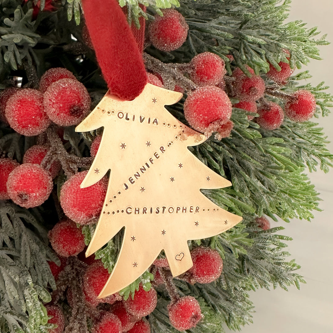 Gold tree-shaped Christmas ornament stamped with names and tiny star details, hanging from a red velvet ribbon on a frosted wreath with red winter berries.