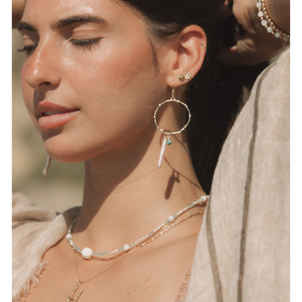 Woman wearing the Gold Pebble Nugget Stud in the ear, shown in warm sunlit tones with layered coastal necklaces and soft natural styling. She is also wearing a gold evil eye stud and pearl and turquoise hoops.
