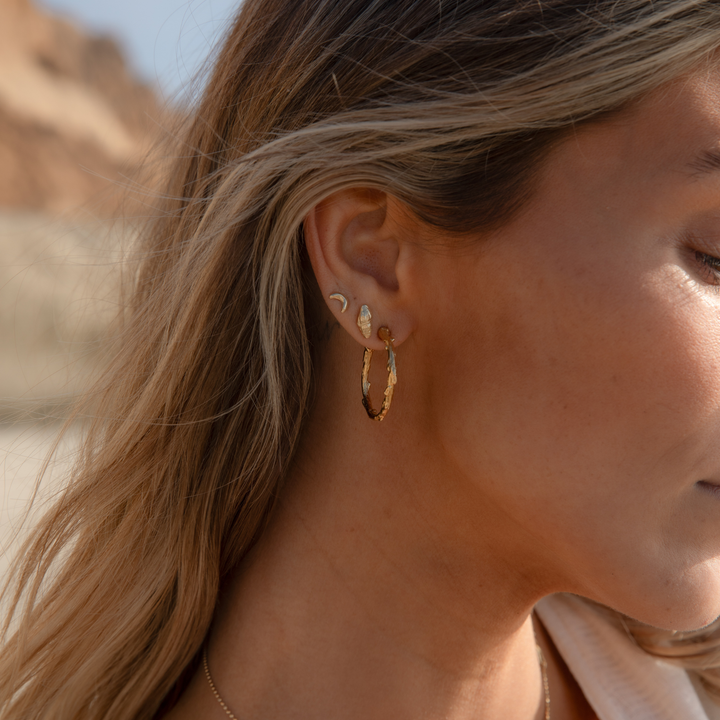 Close-up of a woman wearing textured gold hoop earrings and coastal-inspired seashell and luna crescent moon studs, captured in soft sunlight on a beach.