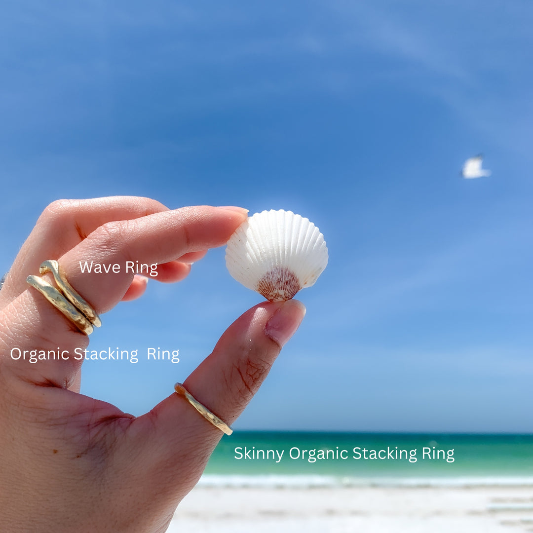 labeled image showing the 14k Wave Ring stacked with the Organic Stacking Ring and Skinny Organic Stacking Ring on a hand holding a seashell at the beach. 