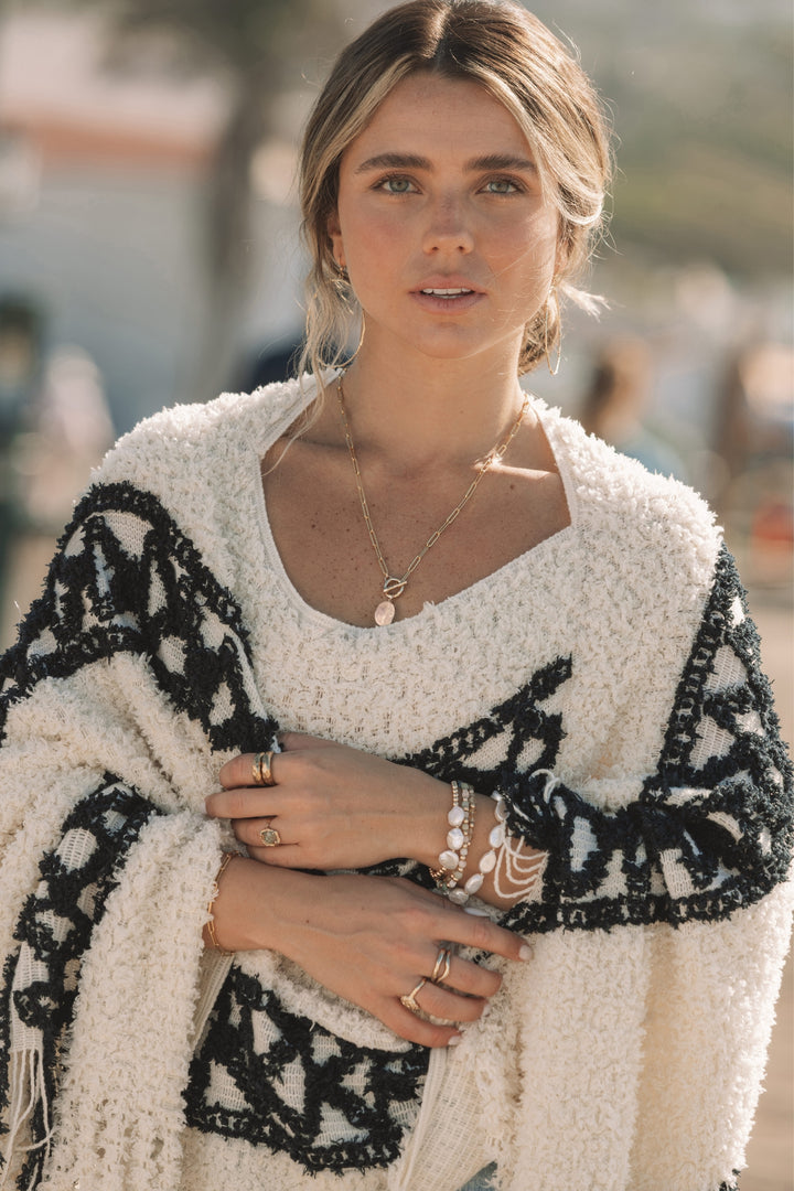 Woman wearing Blooming Lotus Jewelry’s Open Heart Toggle Necklace featuring a faceted morganite gemstone on a hand-carved open heart toggle clasp and gold paperclip chain. Styled with gold rings, pearl and gemstone bracelets, and a cozy boho wrap, photographed in warm coastal sunlight for a soft, natural look.